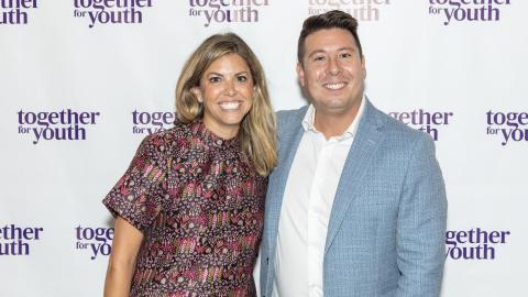 From left to right, a woman and man stand and smile at the camera. They are in front of a white backdrop with purple words reading Together for Youth.